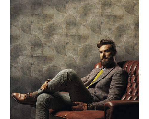 Un bărbat stă așezat în fața unui perete cu tapet cu model circular pe o canapea de piele