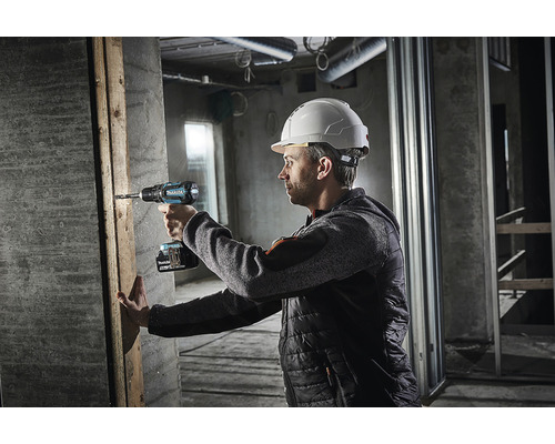 Un bărbat cu cască de protecție găurește cu o șurubelniță electrică o scândură de lemn pe un șantier.