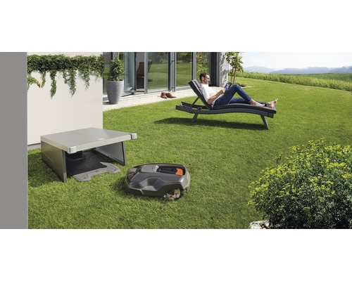Robot de tuns iarba cu stație de încărcare și garaj în grădină, o persoană se relaxează pe un șezlong
