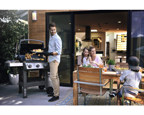 Familie care face grătar afară cu un grătar pe gaz și o masă de sufragerie din lemn.