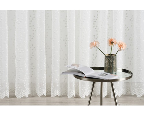 Perdea decorativă albă cu model floral, completată de o măsuță laterală cu o carte și o vază cu flori.