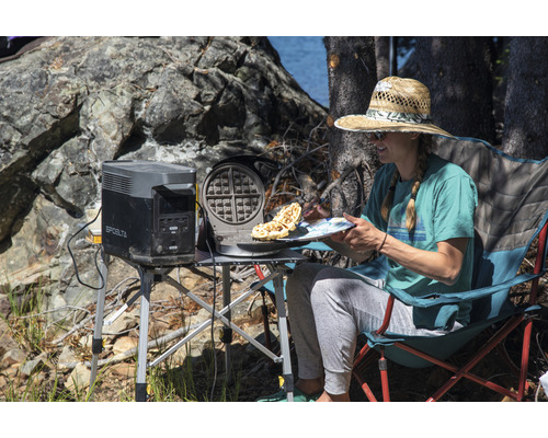 Femeie stând pe un scaun de camping cu un aparat de făcut vafe și o centrală electrică pe o masă afară.