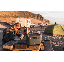 Scenă cu o persoană stând la o masă de picnic lângă o centrală electrică EcoFlow River Pro, cu echipament de camping în fundal.