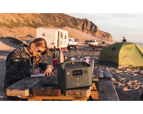 Scenă cu o persoană stând la o masă de picnic lângă o centrală electrică EcoFlow River Pro, cu echipament de camping în fundal.
