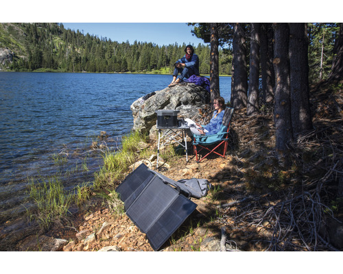 Scenă la lac cu panou solar portabil, stație de alimentare, masă și scaune de camping pentru utilizare în aer liber.