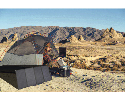 Bărbat lucrează în cort cu laptop, generator de curent și modul solar în peisaj montan.