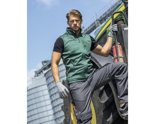 Un bărbat în haine de lucru pozează lângă un tractor. El poartă o vestă și pantaloni de lucru.