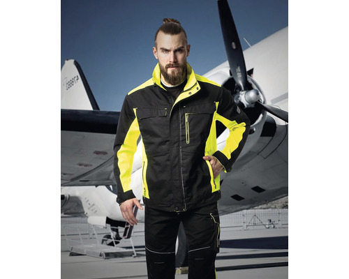 Bărbat în jachetă și pantaloni de lucru negri și galbeni în fața unui avion