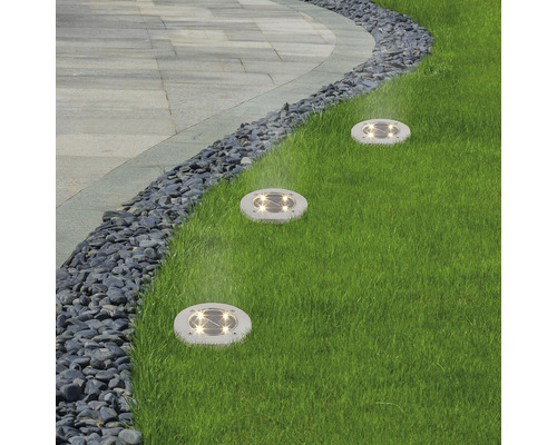 Trei lămpi solare rotunde, la nivelul solului, în gazon, de-a lungul unei căi delimitate cu pietre.