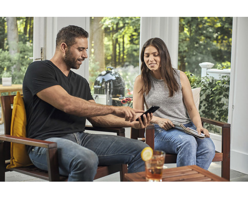 Un bărbat și o femeie stau pe terasă și se uită la un telefon mobil, cu un grătar sferic în fundal.