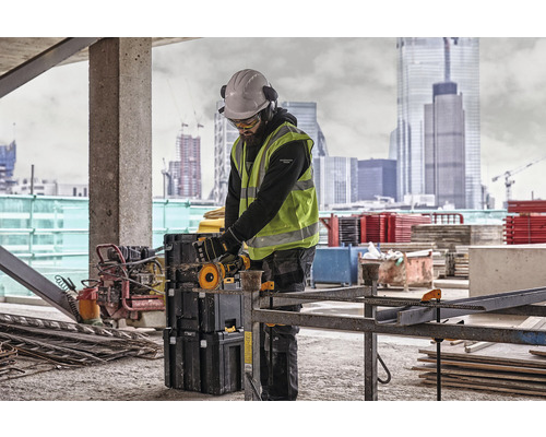 Un muncitor în construcții cu cască de protecție, ochelari de protecție, protecție auditivă și vestă de siguranță lucrează cu un ferăstrău de debitat pe un șantier.
