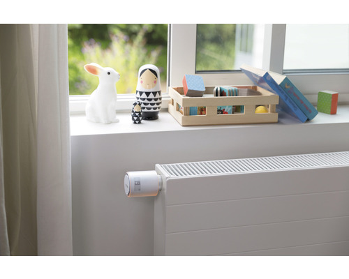 Scenă cu un termostat de radiator pe un radiator într-o cameră de copii cu jucării pe pervazul ferestrei