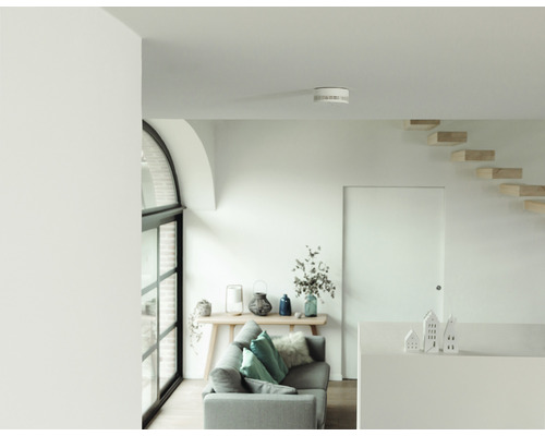 Cameră de zi luminoasă cu detector de fum pe tavan, canapea, masă și scări