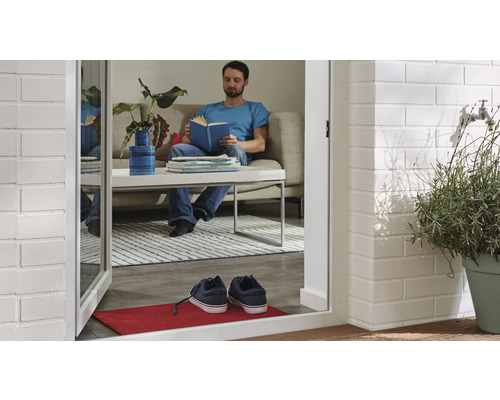 Un bărbat citește o carte într-un living, vizibil printr-o ușă deschisă cu o covorașă și pantofi.