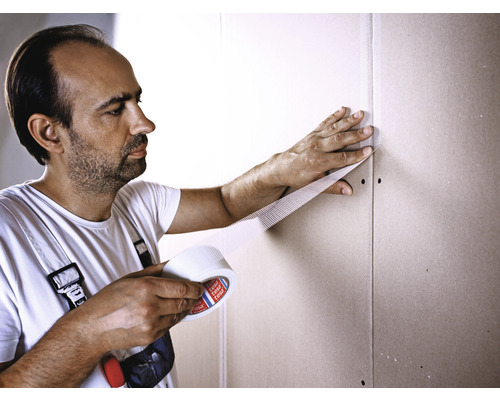 Bărbat care folosește bandă adezivă de armare pe o placă de gips-carton.