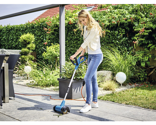 Femeie folosind o perie pentru rosturi pe o terasă pentru a îndepărta buruienile.