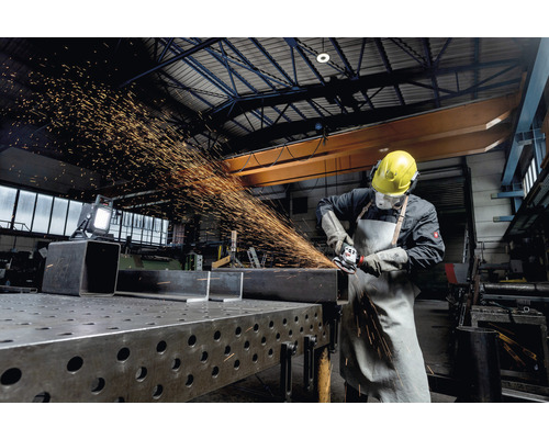 Un muncitor cu îmbrăcăminte de protecție șlefuiește metal cu un polizor unghiular într-un atelier.