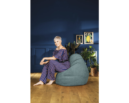 Femeie stând într-o cameră pe un sac de fasole, înconjurată de plante de interior și tablouri.