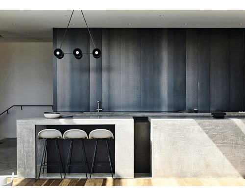 Fotografie de interior a unei bucătării moderne cu o insulă de bucătărie din beton, trei scaune de bar și o lampă suspendată cu trei corpuri de iluminat