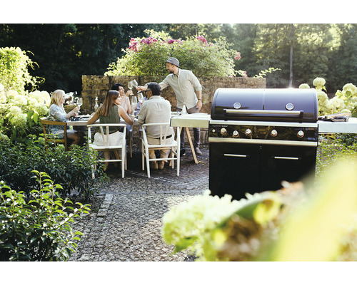 O petrecere cu grătar în grădină cu un grătar pe gaz și un grup de oameni așezați la o masă.