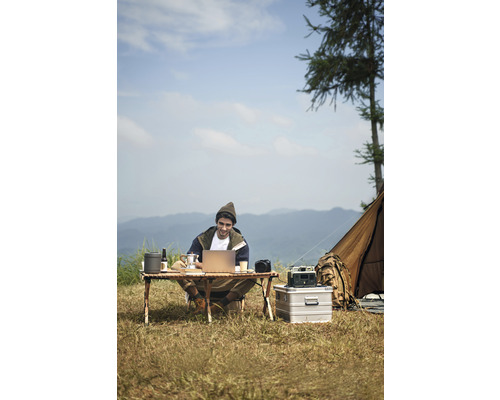 Bărbat stă la o masă de camping și lucrează pe un laptop în aer liber, cu un cort în fundal.