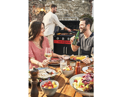 Oameni care fac grătar și mănâncă la o masă de lemn cu un grătar pe gaz în fundal