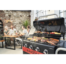 Grătar pe gaz care gătește carne și legume, cu prietenii în fundal