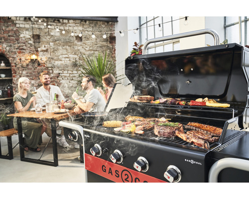 Grătar pe gaz care gătește carne și legume, cu prietenii în fundal