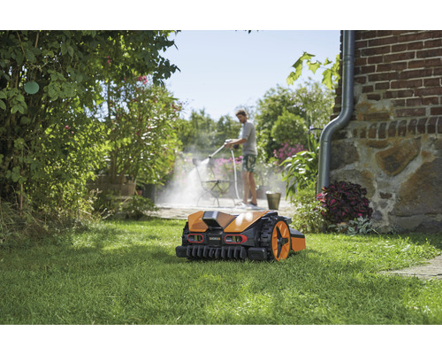 Robot de tuns iarba pe gazon, în fundal o persoană curăță o terasă cu un aparat de curățat cu înaltă presiune