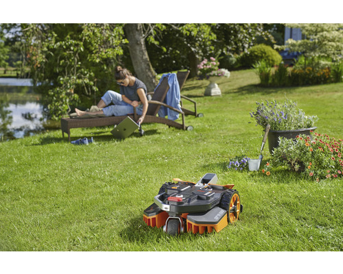 Scenă cu un robot de tuns iarba pe un gazon verde, în timp ce o femeie se relaxează pe un șezlong