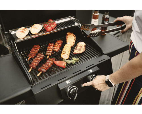 Grătar pe gaz cu produse la grătar și o persoană cu clești de grătar