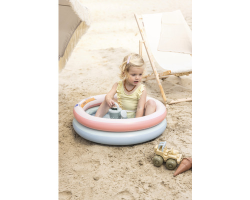 O fetiță stă într-o piscină gonflabilă pentru copii pe plajă, cu jucării de nisip lângă ea