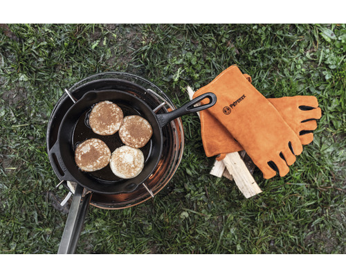 Clătite într-o tigaie din fontă pe un suport de gătit cu mănuși de piele cu logo Petromax lângă lemn.