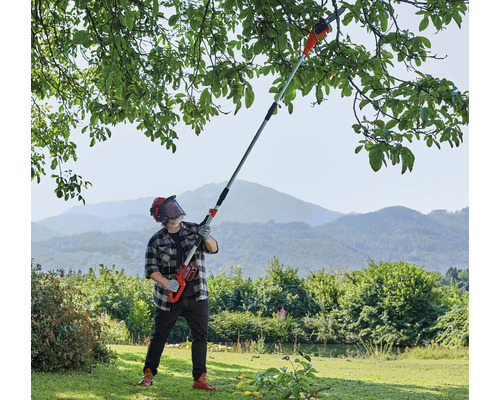 Bărbat cu cască tăind ramuri de copac cu un fierăstrău telescopic cu acumulator în grădină.