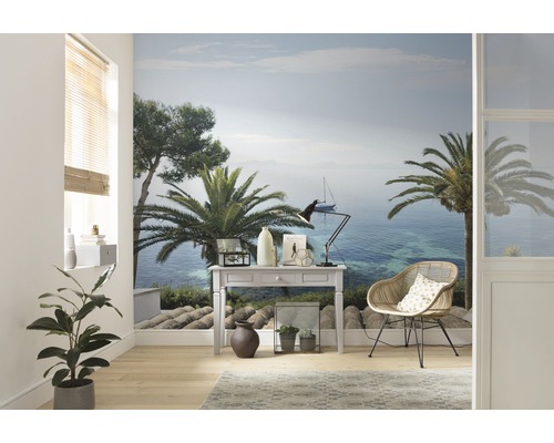 Cameră de zi cu fototapet care prezintă un peisaj de coastă cu palmieri și o barcă cu pânze, mobilată cu un birou și un scaun de răchită.