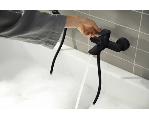 O persoană manevrează o baterie de baie neagră, din care curge apă într-o cadă plină cu spumă.