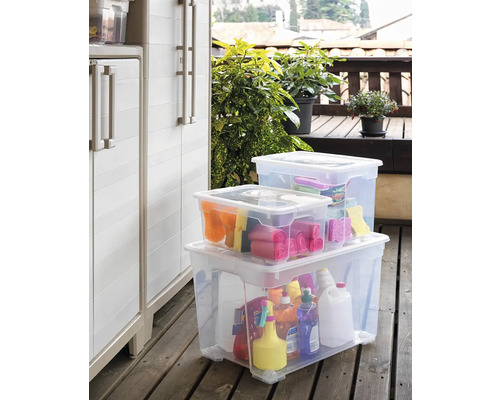 Scenă cu trei cutii de depozitare transparente pe o terasă din lemn, umplute cu diverse obiecte și produse de curățenie, lângă un dulap de depozitare.