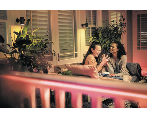 Scenă cu două femei așezate pe o terasă sub lămpi de perete exterioare, uitându-se la un smartphone, înconjurate de plante.