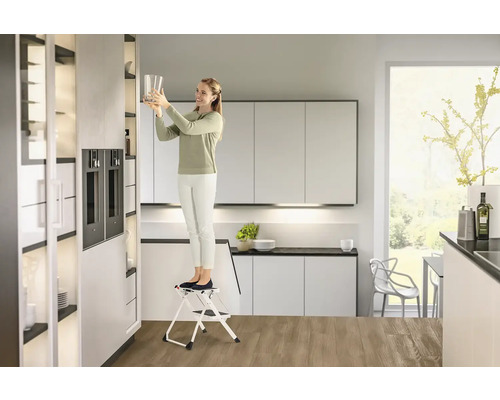 Femeie pe o scară într-o bucătărie modernă punând un pahar într-un dulap