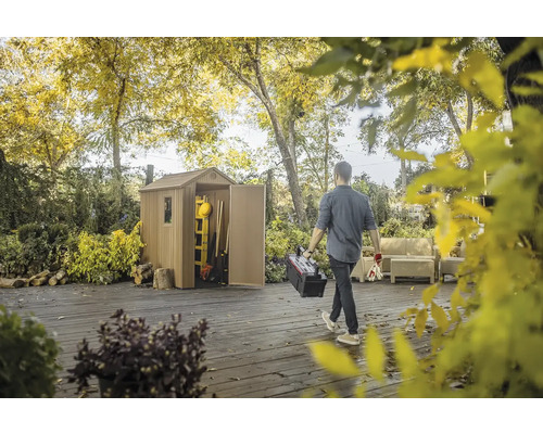 Scenă cu o magazie de grădină în grădină și o persoană care poartă o cutie de scule.