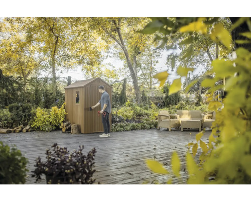 Un bărbat deschide o magazie de grădină înconjurată de copaci și mobilier de grădină.