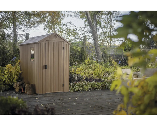 Căsuță de grădină din plastic cu fereastră și ușă pe o terasă de lemn, înconjurată de copaci și plante.