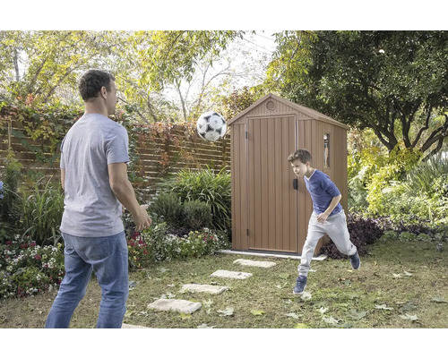 Tată și fiu jucându-se cu o minge de fotbal în fața unei magazii de grădină din grădină.