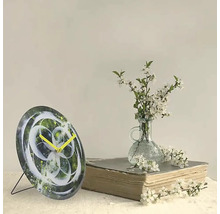 Ceas decorativ de masă cu design abstract și ace galbene lângă o carte și o vază cu flori albe.