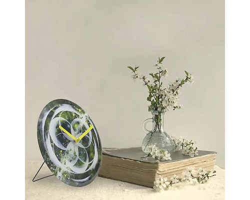 Ceas decorativ de masă cu design abstract și ace galbene lângă o carte și o vază cu flori albe.