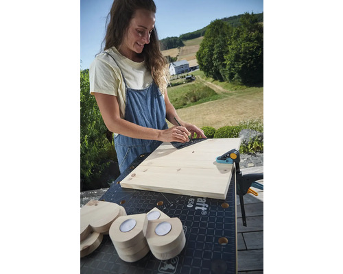 Femeie care trasează o linie pe o scândură de lemn cu un unghi pe bancul de lucru cu o menghină și inimioare din lemn