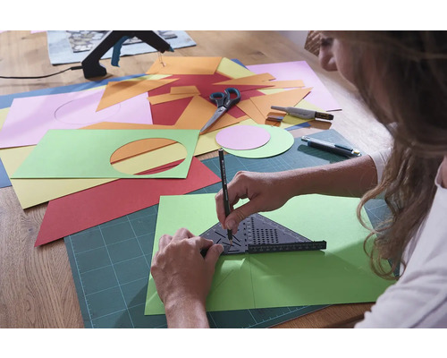 O persoană desenează cu un unghi și un creion pe o foaie verde de hârtie, înconjurată de hârtie colorată, foarfece și un cutter.