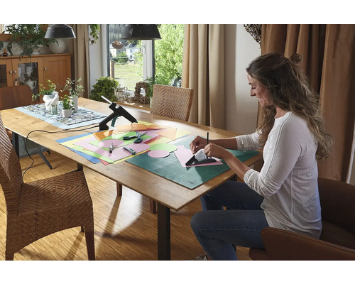 Femeie care meșterește la o masă cu un pistol de lipit, hârtie, foarfece și un suport de tăiat