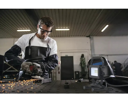 Un bărbat cu ochelari de protecție, șorț și mănuși șlefuiește metal cu un flex, în timp ce un reflector de șantier este în prim-plan.