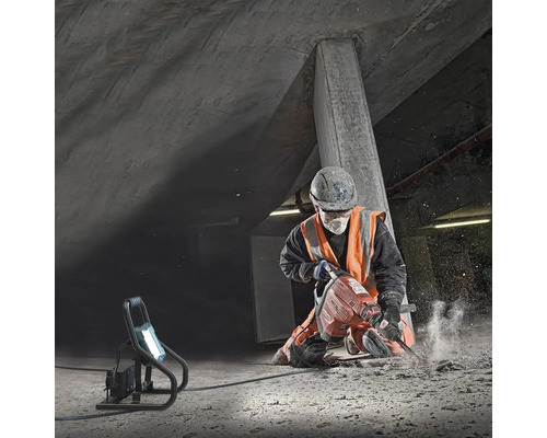 Un muncitor în construcții folosește un ciocan demolator într-o groapă de beton, luminată de o lampă de lucru.